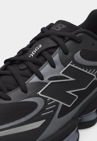 Nærbillede af sort og grå atletiksko med sorte snørebånd, "2000" på tungespidsen og stort "N" logo på siden.