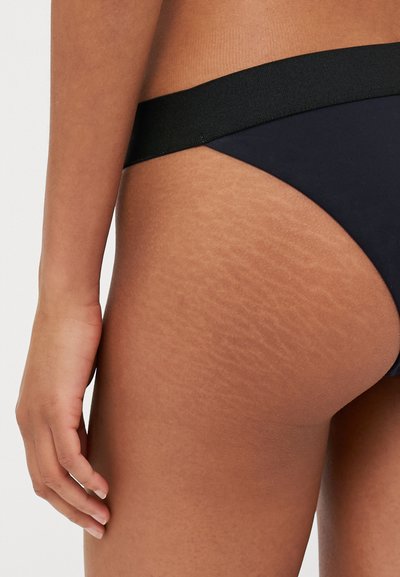 Schwarze Tanga-Unterwäsche mit einem breiten elastischen Bund. Glattes Material mit dezentem Glanz und einstellbarem Schnitt, der die natürliche Körperstruktur zur Geltung bringt.