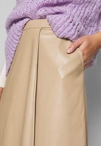 Beige nahkahame, jossa on laskostettu muotoilu, sileä pinta ja näkyvät ompeluyksityiskohdat. Yhdistetty pehmeään laventelinväriseen neulepaitaan.