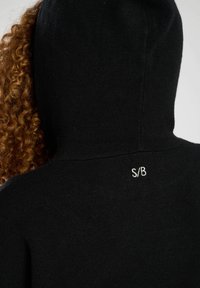 Svart hoodie med en strukturerad tyg, en dragsko huva och en subtil vit "S/B" logotyp broderad på baksidan av nacken.