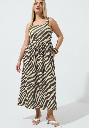 Femme portant une robe midi sans manches à motif zèbre, sandales blanches à talons et bracelets, debout avec une main dans la poche.