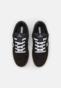 Czarne zamszowe sneakersy z białymi sznurówkami, wyściełanym językiem i podeszwa z gumy w kolorze gumy do żucia. Posiadają metki z brandingiem na języku i boku.