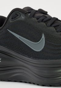 Svart sportskor med en texturerad ovandel, släta mörka detaljer, en framträdande grå Nike-swoosh och en kurvad, tjock sula.