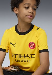 Żółta koszulka piłkarska z czarnymi akcentami, z okrągłym dekoltem, logo Pumy na rękawach oraz herbem Girona FC. Tekst: "ETIHAD AIRWAYS."