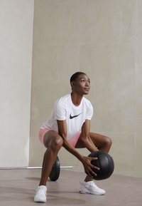 Melnā medicīnas bumba, ko tur abi rokās cilvēks, tērpies baltā Nike krekla un rozā šortos, pietupjas uz gludas grīdas ar neitrālu fonu.