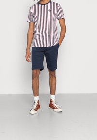 Pánské pruhované tričko s bordovými a bílými vertikálními pruhy, krátkými rukávy, kulatým výstřihem a logem. V kombinaci s námořnickými kraťasy a hnědými vysokými teniskami.