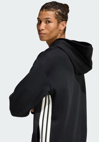 Czarna bluza z kapturem o miękkiej fakturze; posiada trzy białe pionowe paski po każdej stronie; luźny krój, casualowy design.