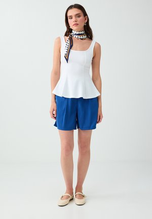 Frau trägt weißes ärmelloses Peplum-Oberteil, blaue knielange Shorts, gepunktetes Halstuch und beige flache Schuhe und steht vor einem einfarbigen Hintergrund.