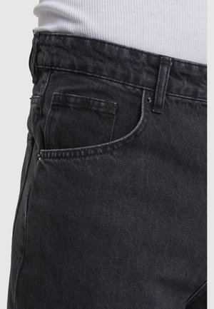 Fekete farmernadrág látható első zsebbel és övbújtatóval, fehér bordázott inggel viselve, amely a deréknál be van tűrve.