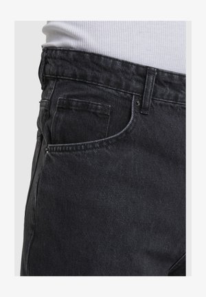 Fekete farmernadrág látható első zsebbel és övbújtatóval, fehér bordázott inggel viselve, amely a deréknál be van tűrve.