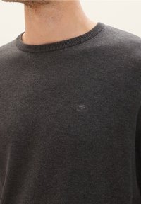 Suéter de punto gris oscuro con cuello redondo y cuello acanalado. Presenta un logo sutil en el frente. Textura suave con un diseño ceñido.