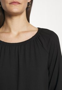 Kvinna som bär en svart top med vid hals och guldiga hoopörhängen, visad från halsen till övre bröstkorgen mot en enfärgad bakgrund.