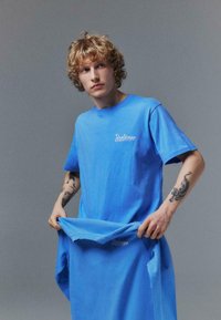 T-shirt bleu à manches courtes en coton, avec un logo imprimé sur la poitrine. Le tissu semble doux et a une texture lisse.