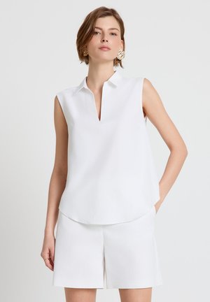 Femme aux cheveux courts portant des boucles d'oreilles en or, un haut blanc sans manches à col, et un short blanc, debout devant un fond clair uni.