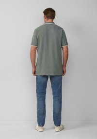 Man staat met zijn gezicht naar een muur, gekleed in een grijs T-shirt met korte mouwen, blauwe jeans en witte sneakers met gestreepte sokken.