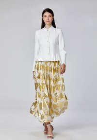Veste blanche à col montant et fermeture à boutons, associée à une jupe maxi fluide à motifs dorés. Des sandales blanches à brides complètent le look.