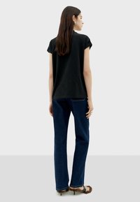 Zwarte t-shirt met korte mouwen, gecombineerd met blauwe straight-leg jeans. Model draagt sandalen met bontaccenten en een zilveren ring, gezien vanuit de rug.