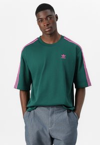Zielona koszulka na krótki rękaw z bawełny, z różowymi trzema paskami na ramionach i małym różowym logo Adidas na piersi.