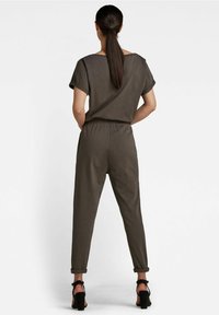 Olijfgroene jumpsuit met korte mouwen, elastische tailleband en opgerolde manchetten. Gecombineerd met zwarte sandalen met hoge hakken, gezien van achteren.
