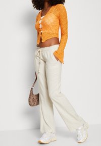 Femme portant un cardigan court en dentelle orange, un pantalon beige à cordon, des baskets blanches, tenant un sac à main en cuir imprimé serpent marron.