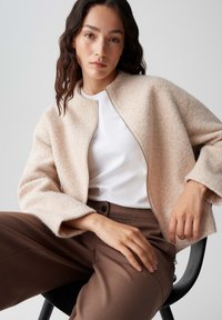 Una giacca di lana texturizzata di un rosa chiaro, con zip frontale e maniche ampie, abbinata a una camicia bianca e pantaloni marroni su misura.