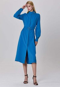 Robe bleue à manches longues avec un col haut, taille cintrée et ceinture avec une boucle en argent. Tissu lisse, longueur genou avec une fente à l'avant.