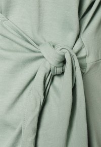 Tecido de malha verde menta com um detalhe de nó torcido de um lado, com textura suave e ligeira elasticidade, exibindo costura limpa.