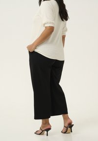 Femme portant un haut blanc texturé à manches courtes, un pantalon noir à jambes larges et des sandales à talons noires, se tenant avec une main dans la poche.