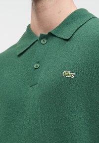 Zaļš polo krekls, izgatavots no teksturētas adītas auduma, ar apkakli un divām pogām, ar mazu Lacoste krokodila logotipu uz krūtīm.