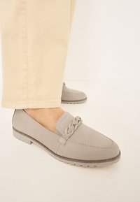 Grå semsket loafers med flettet detalj på toppen, flat gummisåle og en glatt overflate, sammen med lyse beige bukser.