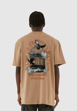 Tan oversized cotton t-shirt with graphic print of a birdcage and birds, featuring text in red and black. Short sleeves and relaxed fit.