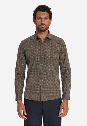 Mann mit kurzen lockigen Haaren und Bart, der ein braunes langärmliges Hemd mit kleinem geometrischem Muster und dunkle Hose trägt.