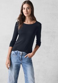 Schwarzes Langarmoberteil mit rundem Halsausschnitt, aus glattem Stoff gefertigt, kombiniert mit hellblauen Jeans mit Knopfverschluss und Fronttaschen.