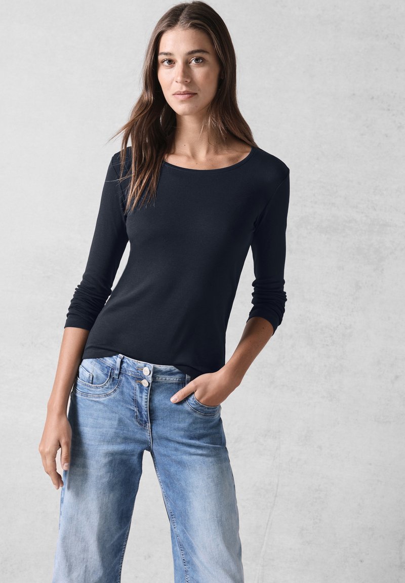 Schwarzes Langarmoberteil mit rundem Halsausschnitt, aus glattem Stoff gefertigt, kombiniert mit hellblauen Jeans mit Knopfverschluss und Fronttaschen.