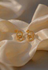 Orecchini in tono oro con un design a cerchio attorcigliato e accenti circolari intrecciati, esposti su un morbido tessuto di satin di colore chiaro.
