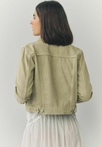 Khaki denim jacket met een aansluitend ontwerp, omgeslagen mouwen en drukknoopsluiting op de taille, gecombineerd met een witte jurk met kantafwerking.