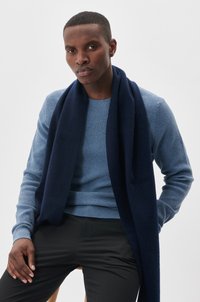 Navy wool scarf draped over a light blue textured knit sweater. Black slim-fit trousers and a neutral background.