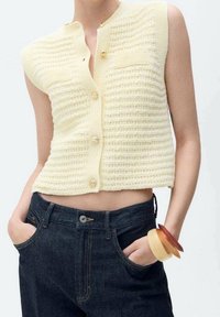 Żółta krótka kamizelka na drutach z teksturowanym wzorem, z przednią kieszenią i czterema guzikami. Połączona z ciemnymi jeansami z wysokim stanem.