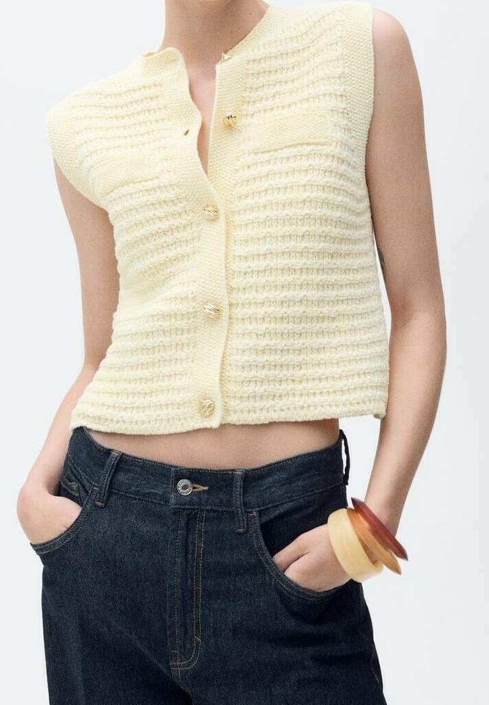 Żółta krótka kamizelka na drutach z teksturowanym wzorem, z przednią kieszenią i czterema guzikami. Połączona z ciemnymi jeansami z wysokim stanem.
