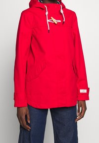 Chaqueta impermeable roja con capucha, cierre de madera con pasador, cordones blancos, bolsillos laterales y un ajuste casual.