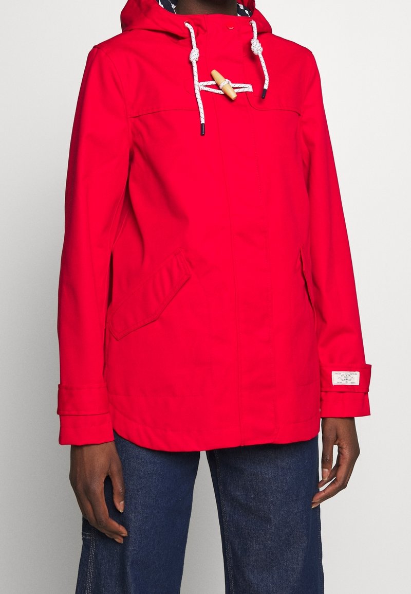 Chaqueta impermeable roja con capucha, cierre de madera con pasador, cordones blancos, bolsillos laterales y un ajuste casual.