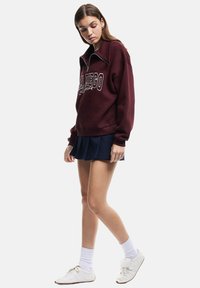 Felpa in pile bordeaux con zip a quarto e stampa bianca "San Diego", abbinata a una gonna plissettata blu navy e scarpe da ginnastica bianche.
