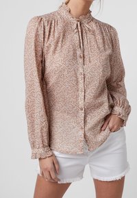 Camicia a maniche lunghe, con bottoni, di colore rosa chiaro con motivo astratto bianco. Presenta un collo arricciato e polsini elasticizzati. Indossata con pantaloni corti bianchi.