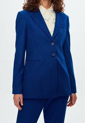 Blazer taillé bleu avec deux boutons, revers crantés et tissu texturé. Présente des poches latérales et une silhouette structurée.