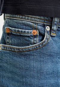 Κοντινή λήψη μπλε τζιν με επίκεντρο την τσέπη για κέρματα, τα χάλκινα καρφιά και τη ραφή αντίθεσης κάτω από ένα σκοτεινό υφασμάτινο πουκάμισο.