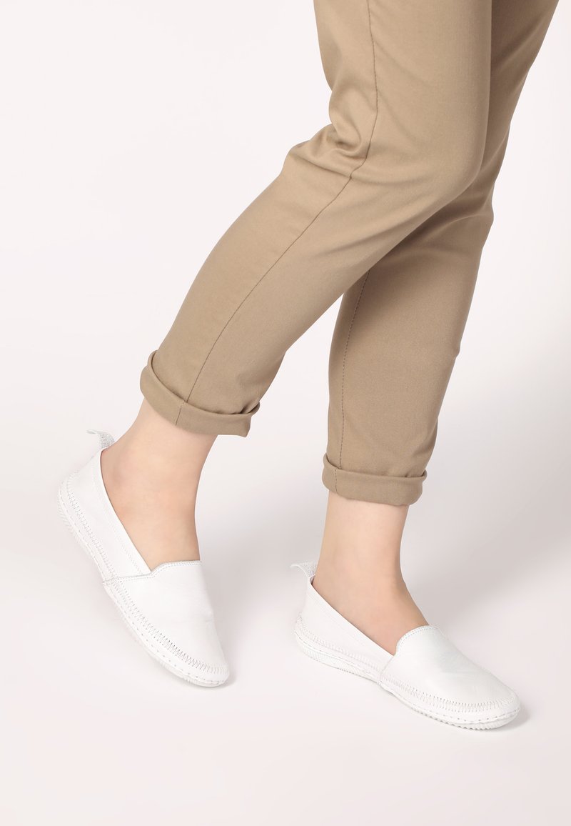 Weiße Slipper aus glattem Leder mit elastischen Öffnungen, ausgestattet mit einem gesteppten Design und einer flexiblen Sohle, kombiniert mit hochgekrempelten beigen Hosen.