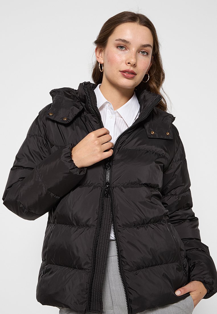 Frau in einer schwarzen Daunenjacke mit hohem Kragen, teilweise zugezippt über einem weißen Hemd, posiert mit einer Hand am Reißverschluss und der anderen in der Tasche.