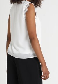 Top blanc sans manches avec des bordures en dentelle sur les épaules, texture lisse et ourlet inférieur légèrement transparent. Associé à un pantalon noir.