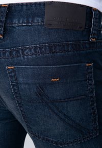 Vaqueros de mezclilla azul oscuro con acentos de costura en naranja, etiqueta de marca de cuero y diseños detallados en los bolsillos. Vista de cerca de la textura de la tela.