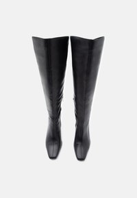 Μαύρες δερμάτινες μπότες έως το γόνατο με μυτερές μύτες και πλευρικά φερμουάρ, όρθιες σε απλό λευκό φόντο.